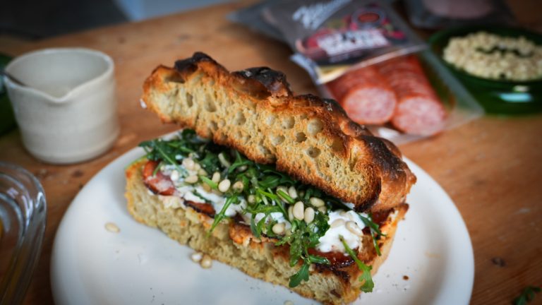 Focacciamacka med burrata, ruccola, pinjenötter och het chilisalami liggandes på ett vitt fat.